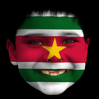 A lovely cheerful six year old boy smiling with broken incisors and half grown incisor teeth with Suriname flag painted on his face. Horizontal bands of green, white, red,white, and green. There is a large, yellow, five-pointed star in the center of the red band.