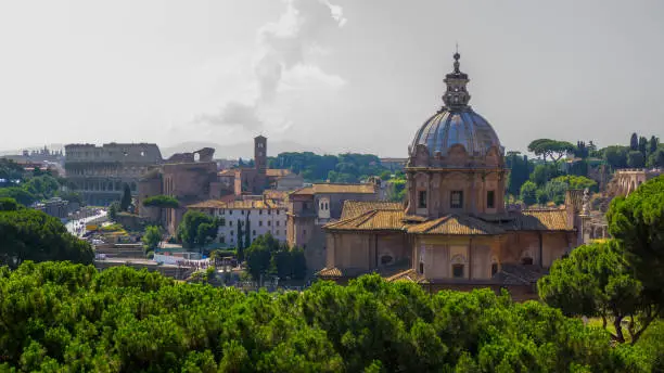 Historical attractions and old architecture of Rome: the Colosseum, the Church of Santi Luca e Martin, Basilica of Cosmas and Damian, the ancient ruins of the Forum of Julius Caesar, Temple of Peace – photo from the terrace of the building of the Forum with a beautiful sky in shades of grey and the hills on the horizon and green trees
