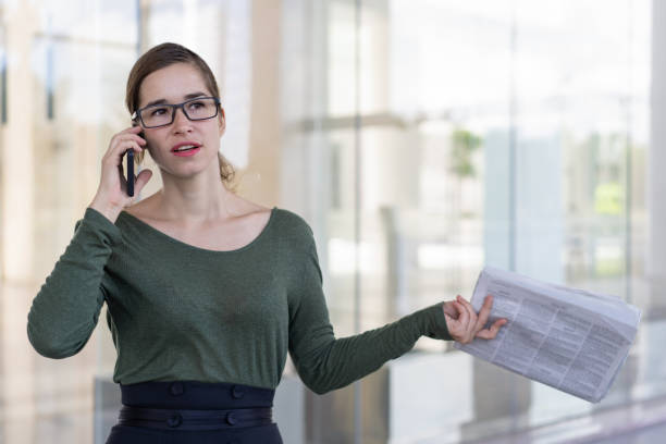 Serious business expert concerned with latest news and making phone call. Young woman in glasses holding newspaper while talking on cell outdoors. Bad news concept