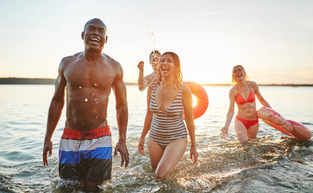 смеющиеся молодые друзья в купальниках веселились в озере - inner tube swimming lake water стоковые фото и изображения