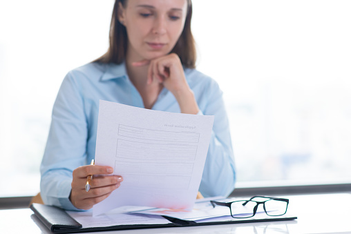 Serious woman reading and examining document. Entrepreneur sitting at desk and working. Paperwork and recruitment concept. Front view.