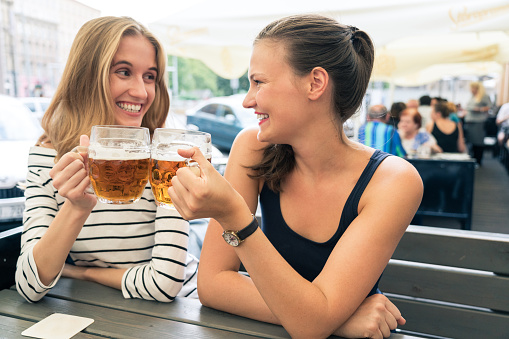 Portrait of two young woman friends sitting at outdoors cafe and toasting beers