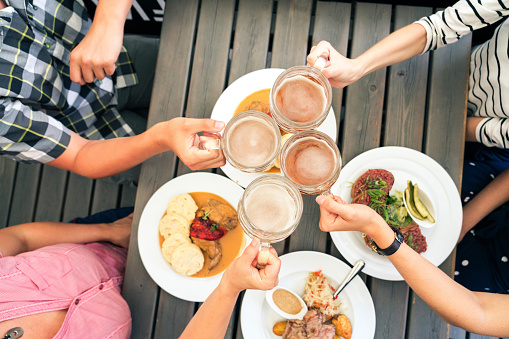 Top view of hands of friends clinking big pints with beer with snacks on the table. Four friends toasting beer glasses in a cafe.