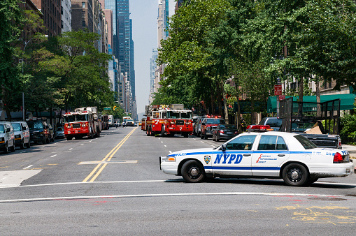 Several cars, trucks, ladders and firefighters of the New York Fire Department at work in East 57th Street near Sutton Place. The street is blocked by a patrol car of the New York Police Department.