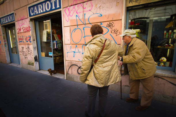 Rome, Italy: Two Seniors Walk in San Lorenzo Neighborhood Rome, Italy: Two seniors—a woman and a man with a cane—walk in front of a pink wall near a shop in the San Lorenzo neighborhood of Rome. san lorenzo rome photos stock pictures, royalty-free photos & images