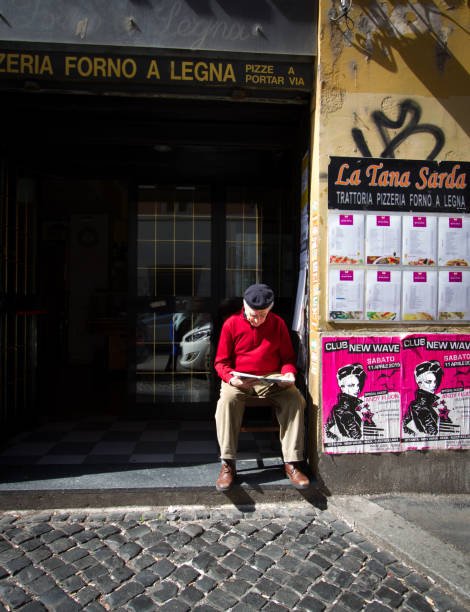 Rome, Italy: Beret-Wearing Senior Reads in Sunlit Doorway Rome, Italy: A beret-wearing senior man reads a newspaper in a sunlit doorway in the graffiti-covered San Lorenzo neighborhood of Rome. san lorenzo rome photos stock pictures, royalty-free photos & images