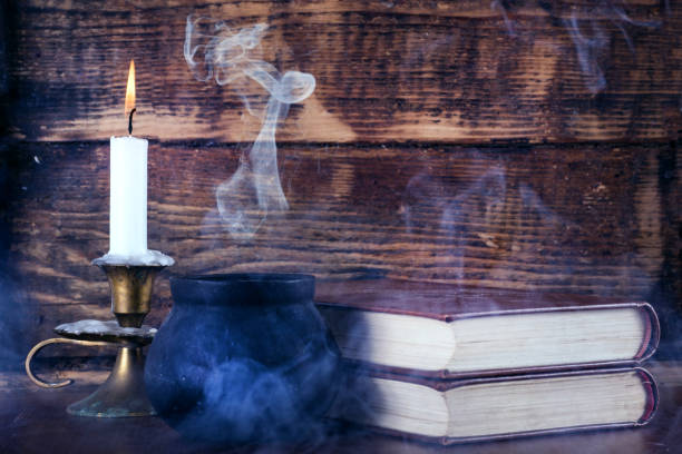Old books of magic and witch pot with smoke and candle Books of magic and small black witch pot with smoke and wax candle burning in candlestick. Old wooden background. Halloween concept. Close up, selective focus cauldron photos stock pictures, royalty-free photos & images