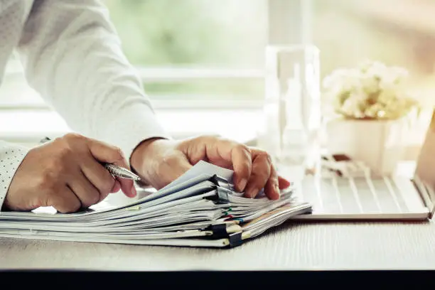 Businessman hands holding pen for working in Stacks of paper files searching information business report papers and piles of unfinished documents achieves on laptop computer desk in modern office