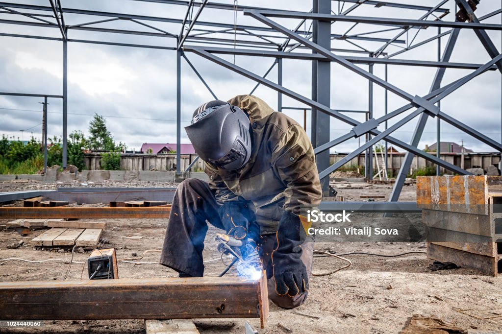 A young  man welder A young  man welder in brown uniform, welding mask and welders leathers, weld  metal  with a arc welding machine at the construction site, blue sparks fly to the sides Welder Stock Photo