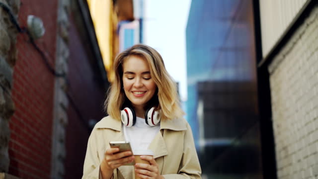 Slow motion of happy girl using smartphone while walking in the street wearing coat and holding take-away coffee. Urban lifestyle, millennials and young people concept.