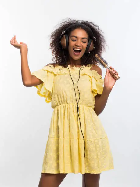 Young pretty african american woman in yellow dress singing and dancing with hair dryer instead microphone at white wall. Black girl having fun isolated on background