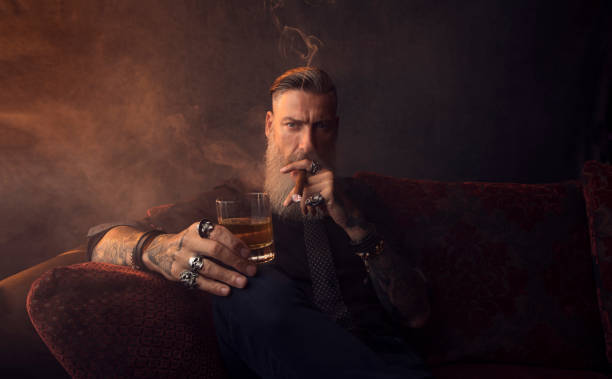 Portrait of an attractive business man with a cigar and a glass of whiskey in a dark room Portrait of an attractive business man with a cigar and a glass of whiskey in a dark room man cigar stock pictures, royalty-free photos & images