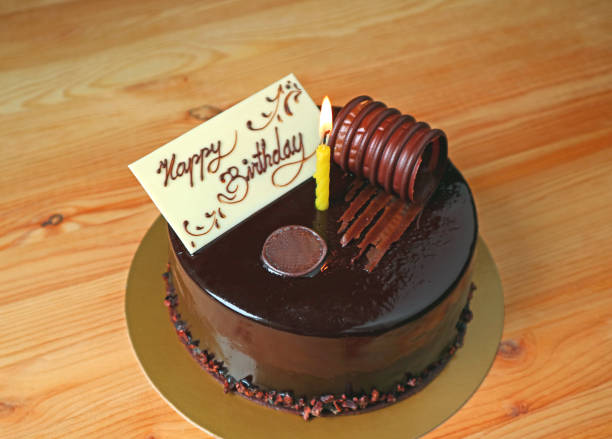 istock Chocolate Mousse Birthday Cake Topped with White Chocolate Greeting Card and a Lighting Candle on the wooden table 1024842876