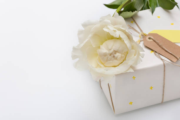 Nice present packed in white paper with small yellow stars with beautiful peony flower on white wooden background. Wrapped gift for birthday party.   Mock-up. Holidays concept.
