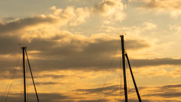 Part view of two top masts silhouettes of sailboats in foreground during a sundown with a yellowish sky with many clouds in the background
