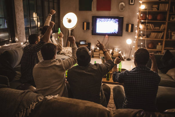 Friends spend weekend together watching TV Group of young men gather at home party. They are watching basketball game on tv, drinking beer, making jokes and cheer for their team. watching stock pictures, royalty-free photos & images