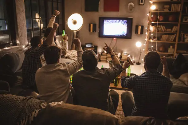 Group of young men gather at home party. They are watching basketball game on tv, drinking beer, making jokes and cheer for their team.