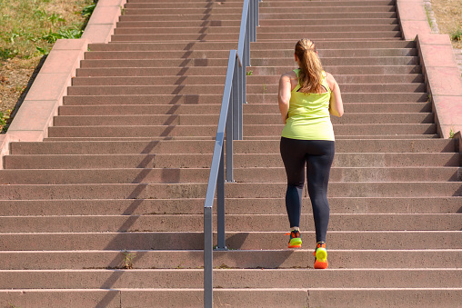 Full length rear view of an active and determined middle-aged woman running while climbing stairs during intense workout for weight loss outdoors in a sunny day
