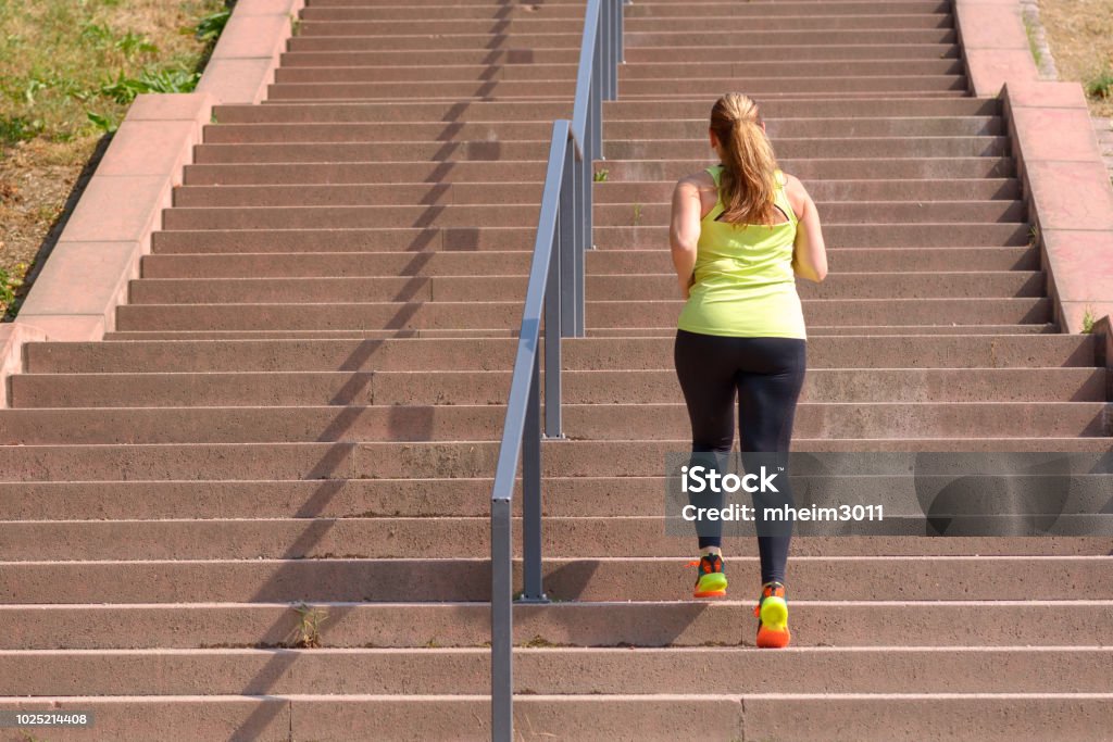 Woman running while climbing stairs during workout Full length rear view of an active and determined middle-aged woman running while climbing stairs during intense workout for weight loss outdoors in a sunny day Exercising Stock Photo