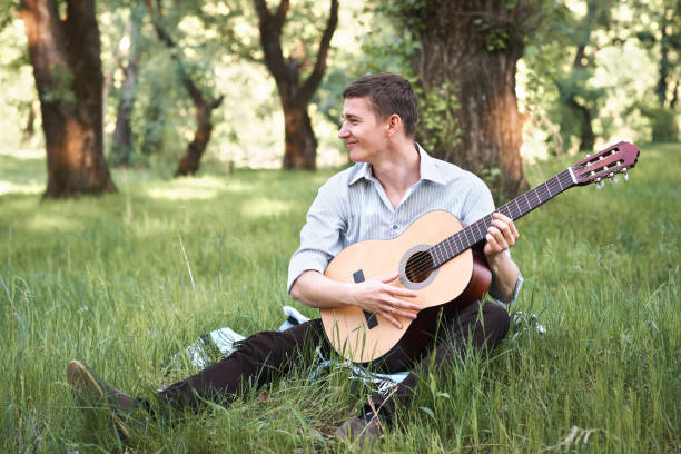 istock young man playing guitar in the forest, sit on the grass, summer nature, bright sunlight, shadows and green leaves, romantic feelings 1025310286