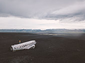 Drone aerial view of Young man stands on airplane crashed on black sand beach looking around contemplating surroundings