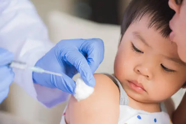 Photo of Doctor giving an injection vaccine to a girl. Little girl crying with her mother on background.