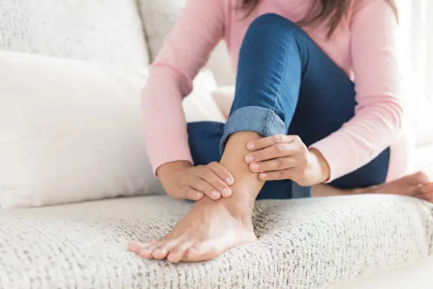 Photo of Closeup woman sitting on sofa holds her ankle injury, feeling pain. Health care and medical concept.