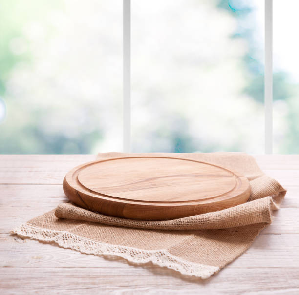 istock Empty textured wooden pizza board with napkin on the table and kitchen window blurred background 1025477544