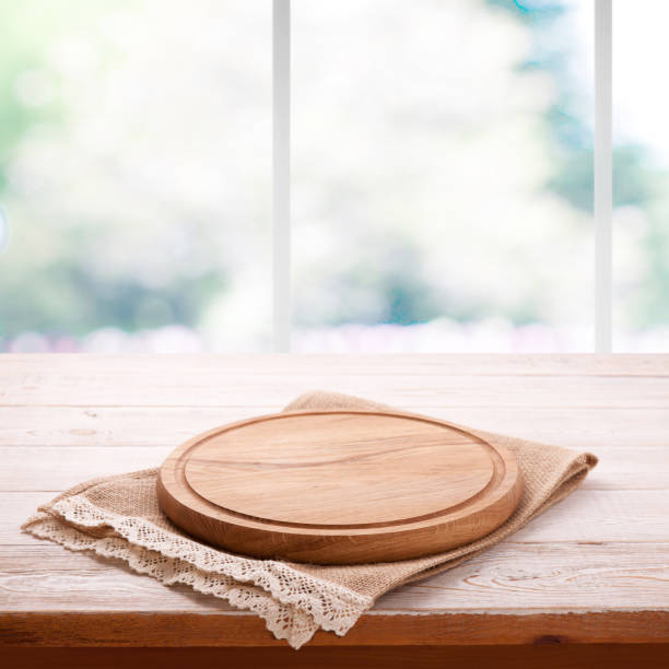 istock Empty textured wooden pizza board with napkin on the table and kitchen window blurred background 1025477552