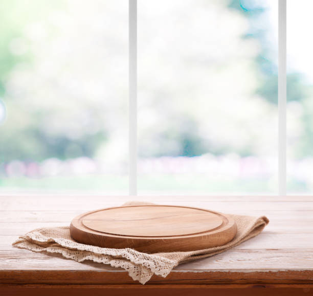 istock Empty textured wooden pizza board with napkin on the table and kitchen window blurred background 1025477572