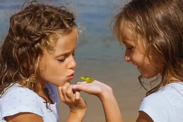 Photo of Twin girls on the beach, one is holding a little baby frog, while the other girl is trying to kiss it