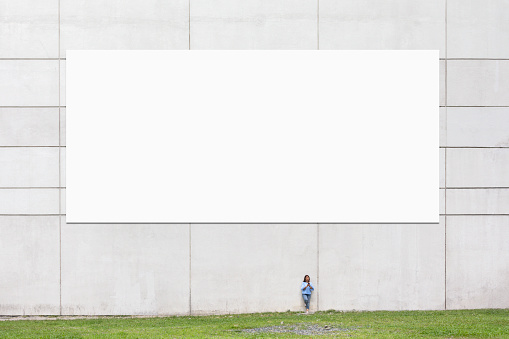 A woman using her phone by a wall with a large blank billboard in a city environment.