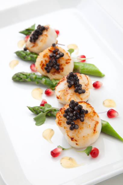 Close up of delicious appetizer scallops with black caviar. Garnished with asparagus, pomegranate seeds, microgreens, and sauce. Elite food.\