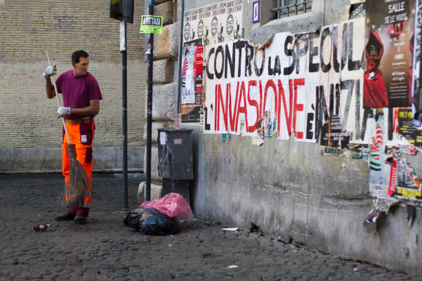 Rome, Italy: A street sweeper in orange pants cleans the cobblestones near a grungy wall and garbage in Rome’s Trastevere neighborhood.