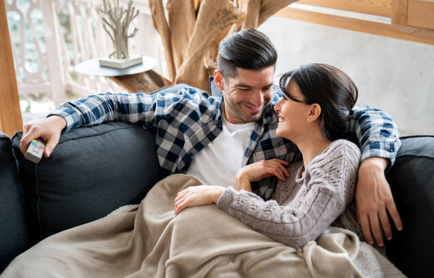 Loving couple watching tv in their winter lodge while lying on the sofa Portrait of a loving couple watching tv in their winter lodge while lying on the sofa looking very happy - lifestyle concepts log cabin photos stock pictures, royalty-free photos & images