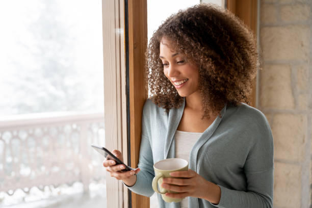 beautiful woman at home drinking a cup of coffee and texting on her phone - window home interior women people imagens e fotografias de stock