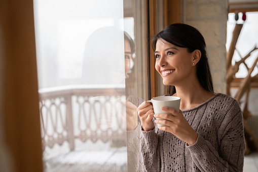Beautiful woman at home drinking a cup of coffee and looking through the window while smiling - lifestyle concepts