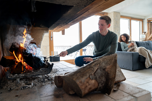 Portrait of a man burning logs in the fireplace while relaxing with his partner at a winter lodge - lifestyle concepts