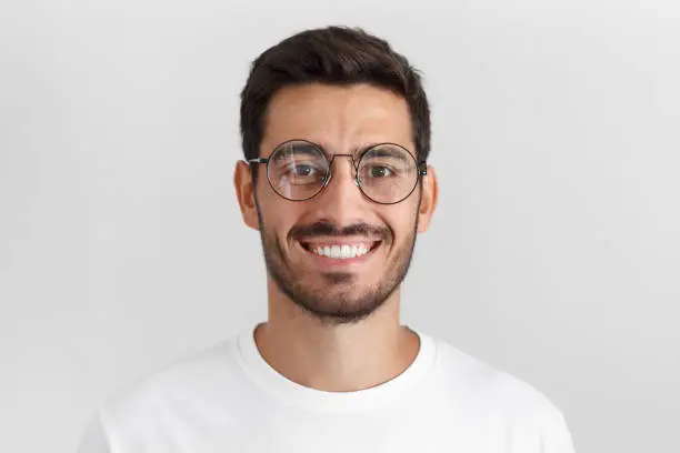 Photo of Daylight portrait of young handsome caucasian man isolated on grey background, dressed in white t-shirt and round eyeglasses, looking at camera and smiling positively