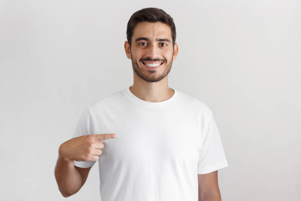 дневной снимок улыбающегося молодого человека, указывающего на его пустую белую футболку с указательным пальцем, копировать пространство  - европейского происхождения стоковые фото и изображения