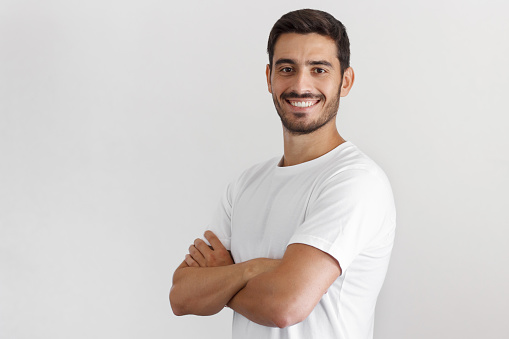 Indoor daylight portrait of young european caucasian man isolated on gray background, standing in white t-shirt with arms crossed, smiling and looking straight at camera