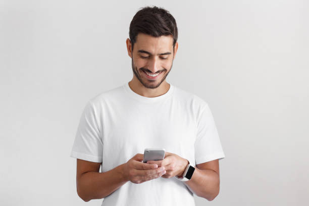 daylight portrait of young european caucasian man isolated on gray background wearing white t-shirt standing in front of camera, looking attentively with smile at screen of smartphone he is holding - attentively imagens e fotografias de stock