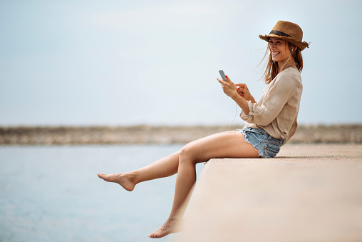 Happy girl enjoying summer holiday sitting on the dock using smart phone and earphones