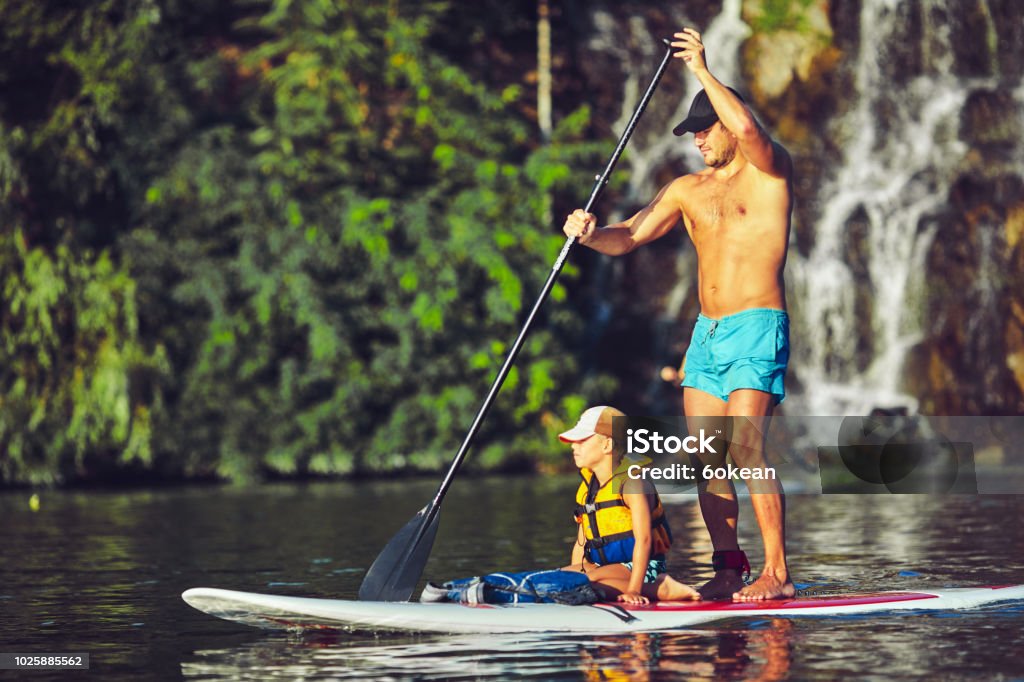 positive smiling boy in rashguard and his young father enjoying stand up paddleboarding Family Having Fun Stand Up Paddling Together in the Ocean on Beautiful Sunny Morning Family Stock Photo