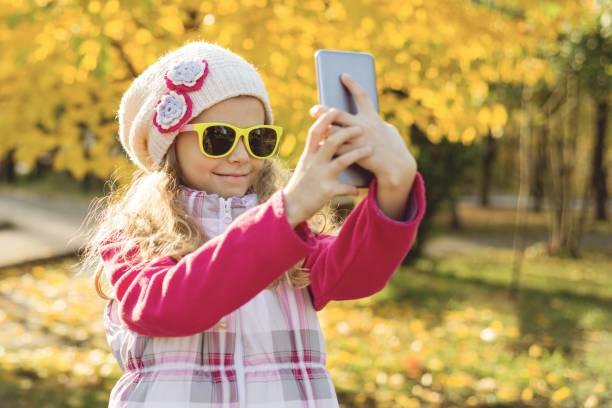 красивая девушка 7 лет делает селфи с помощью смартфона, осенний фон. - 6 7 years lifestyles nature horizontal стоковые фото и изображения