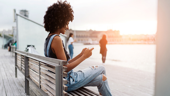 Skilled attractive student female with afro hairstyle wearing jeans overalls is typing messages on a display of a mobile phone while sitting on a bench at the city berth area on a sunset. Flare light