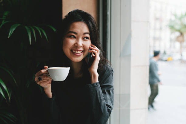 istock Charming asian female freelancer is talking on a phone while sitting in a cafe with beside a big window. Young attractive business woman is making a business call during the lunch break. 1025942376