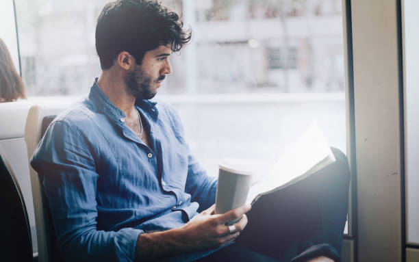 Young bearded office employee male reading newspaper and drinking takeaway coffee while going to the work by public transport to avoid morning traffic jam for cars. Stylish man reading daily news.