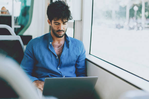 istock Young man is reading emails on portable computer while sitting in a city bus beside a window. Hipster guy with dark curly hair wearing light blue shirt is going to the office by a public transport. 1025958940