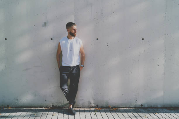 istock Bearded hipster guy wearing a blank white t-shirt is looking aside while standing on a light gray concrete wall background on a street. Horizontal mock up. Empty space for logo or design. 1025963640